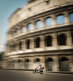 Blurred motion of people riding motor scooter on road against coliseum