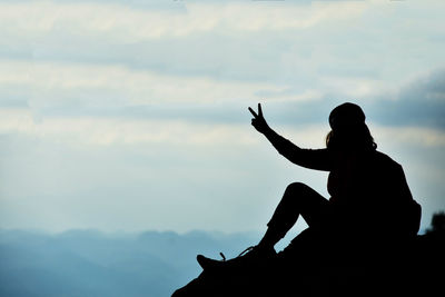 Woman gesturing peace sign while sitting on cliff against sky