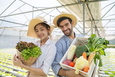 Portrait of smiling man and woman holding basket with vegetable standing at vegetable farm