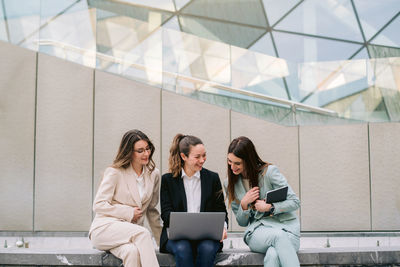 Company of smiling businesswomen in smart casual clothes sitting in city and using laptop while working on startup project together