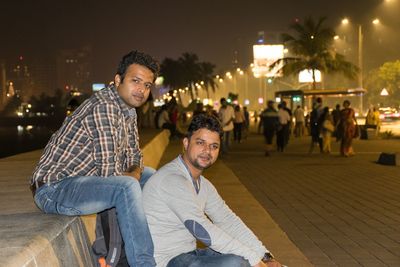 Portrait of smiling young man sitting in city against sky at night