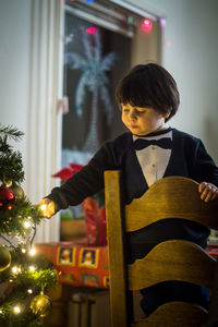 Boy touching illuminated christmas tree while standing on chair at home
