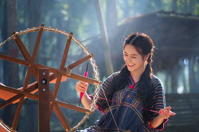 Young woman weaving thread on wheel against house