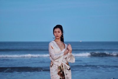 Portrait of young woman standing at beach against sky