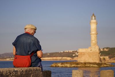 Rear view of senior man sitting on retaining wall with chania lighthouse in background