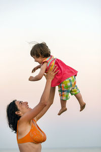 Happy mother playing with daughter at beach against clear sky during sunset