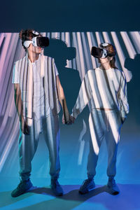 Front view of couple in modern vr goggles holding hands while standing on blue background with glowing lights in studio