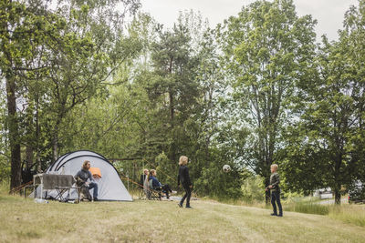 Children playing soccer ball while family sitting by tent at campsite