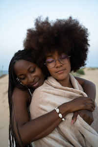 Happy young multiracial female best friends wrapped in warm blanket cuddling with eyes closed while chilling and enjoying time together in summer evening during holidays on seashore