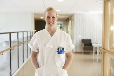 Portrait of happy female nurse standing with hands in pockets at hospital corridor