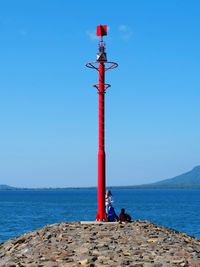 People relaxing by lighthouse on groyne by sea against clear blue sky