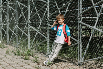 Full length of boy talking on mobile phone while leaning against chainlink fence
