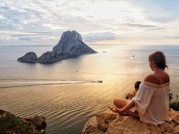 Mother meditating while sitting with daughter on cliff against sea