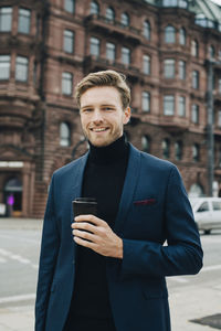 Portrait of businessman with disposable cup standing against building in city