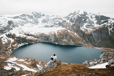 Rear view of man on cliff in front of lake and snowcapped mountains