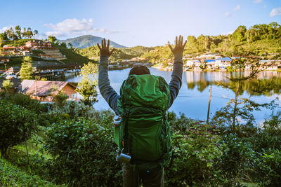 Rear view of man with arms raised standing against lake by village