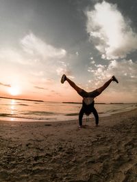 Man doing handstand at beach against sky during sunset