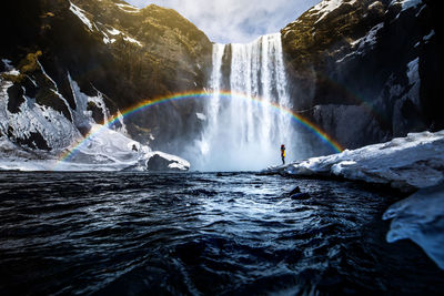 Unrecognizable traveler standing near amazing waterfall and rainbow in mountains during vacation in iceland