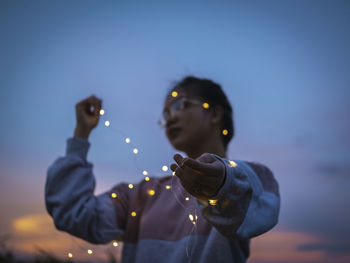 Defocused image of young woman holding string lights standing outdoors