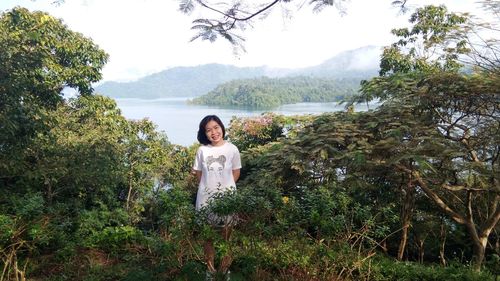 Portrait of woman smiling while standing amidst trees against lake