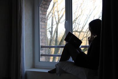 Woman reading book while relaxing on window sill