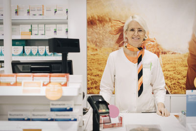 Portrait of confident female pharmacist standing at checkout against poster in medical store