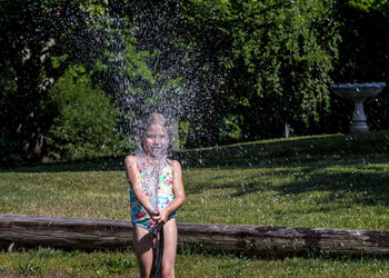 Playful girl in swimsuit spraying water with garden hose at lawn