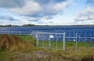 View of solar panel farm with fence on field against sky, germany