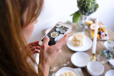 Rear view of woman photographing waffles in plate on table by wall at home