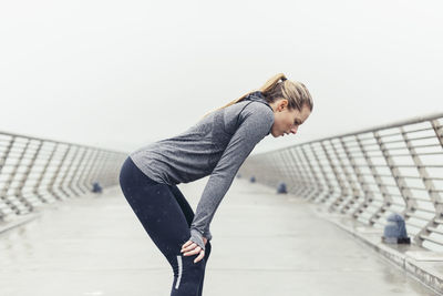 Side view of tired female athlete standing with hands on knees on bridge against clear sky