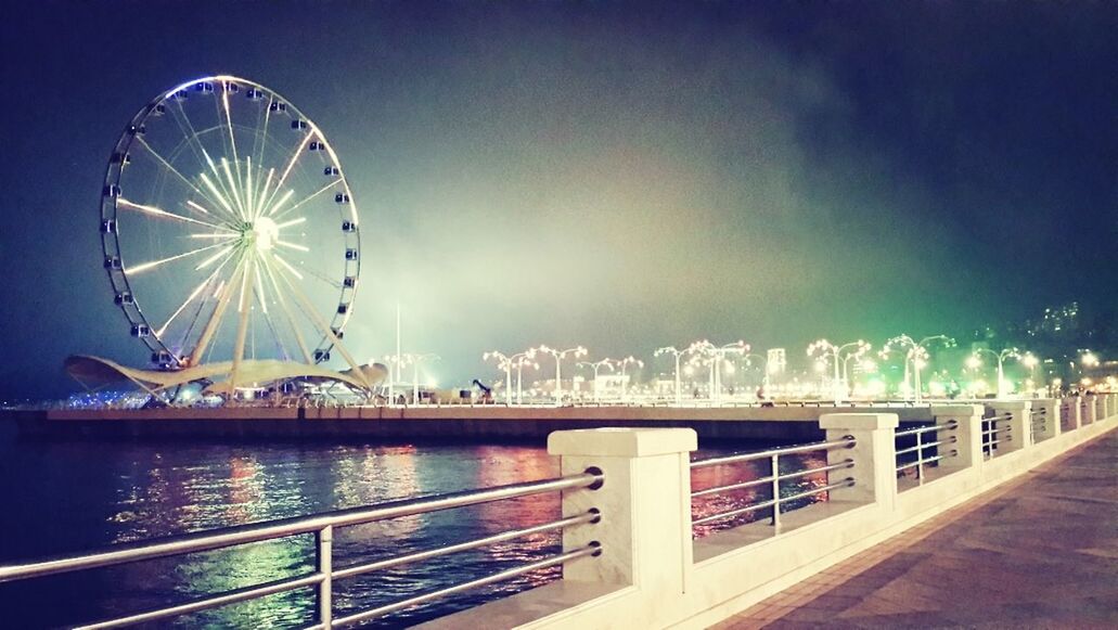 ferris wheel, amusement park, illuminated, amusement park ride, built structure, arts culture and entertainment, architecture, sky, water, night, railing, blue, building exterior, river, bridge - man made structure, city, outdoors, sea, travel destinations, connection