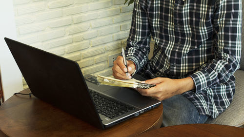 Midsection of businessman writing in book while using laptop 