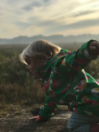 Side view of baby girl kneeling on tree trunk against sky during sunset