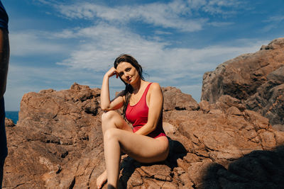 Portrait of sensuous young woman sitting on rock against sky