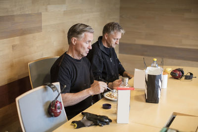 Workers having lunch at table while sitting against wall in industry