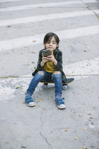 From above full body of adorable boy in casual clothes browsing cellphone while sitting on skateboard on pedestrian crossing in city