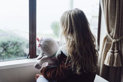 Rear view of girl holding stuffed toy while looking through window