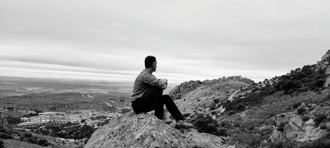 sky, one person, mountain, rock, leisure activity, nature, scenics - nature, cloud, landscape, adult, black and white, environment, beauty in nature, monochrome photography, activity, full length, monochrome, tranquility, men, lifestyles, looking at view, white, tranquil scene, solitude, adventure, non-urban scene, mountain range, vacation, rear view, remote, trip, hiking, holiday, land, outdoors, travel, day, walking, copy space, sitting, idyllic, contemplation, relaxation, travel destinations, young adult, loneliness, recreation, black, exploration