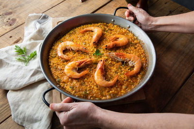 From above crop chef holding large metal pan of while appetizing nutrient paella with roasted shrimps against rustic table in kitchen at home