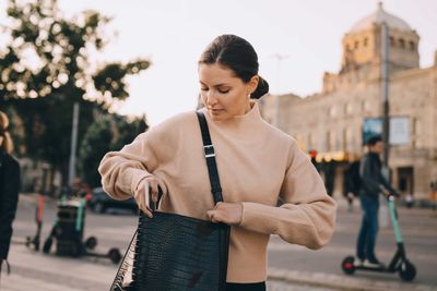 Fashionable woman searching in bag while standing on footpath