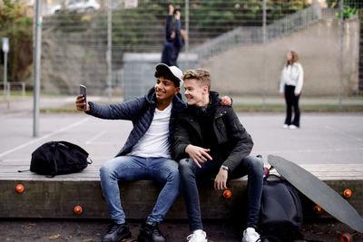 Full length of teenage boys taking selfie with mobile phone while sitting at park