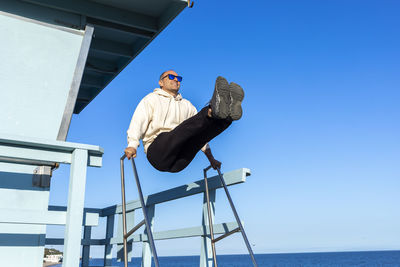 Low angle view of young man standing against sea against clear blue sky
