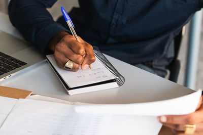 Crop black adult male in formal clothes sitting at table while taking notes in notebook from papers near netbook in bright workplace