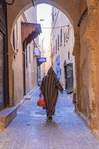 Rear view of man walking in traditional hood clothing while carrying oranges