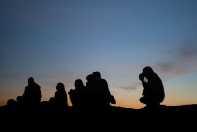 Silhouettes of seated people on the ground at dusk, photographed from behind during beautiful sunset