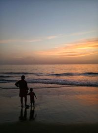 Silhouette father and son standing at beach against sky during sunset