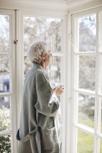 Contemplative senior man holding coffee cup while looking through window at home