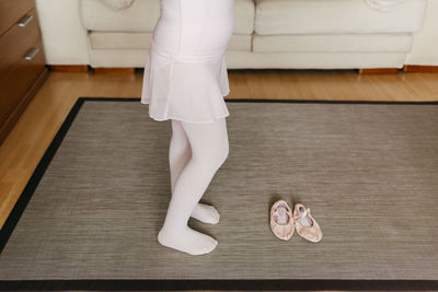 From above anonymous girl in light leotard and tights standing on carpet near dance shoes during training at home