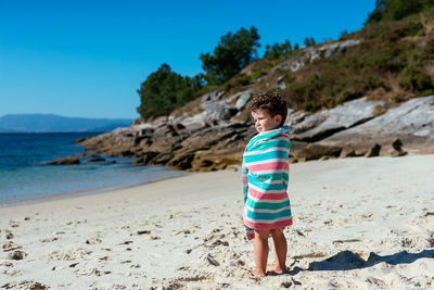 Full length of barefoot boy wrapped in beach towel standing on sandy coast of sea after swimming in sunlight