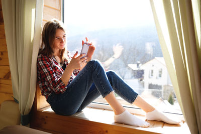 Young woman using phone while sitting on window